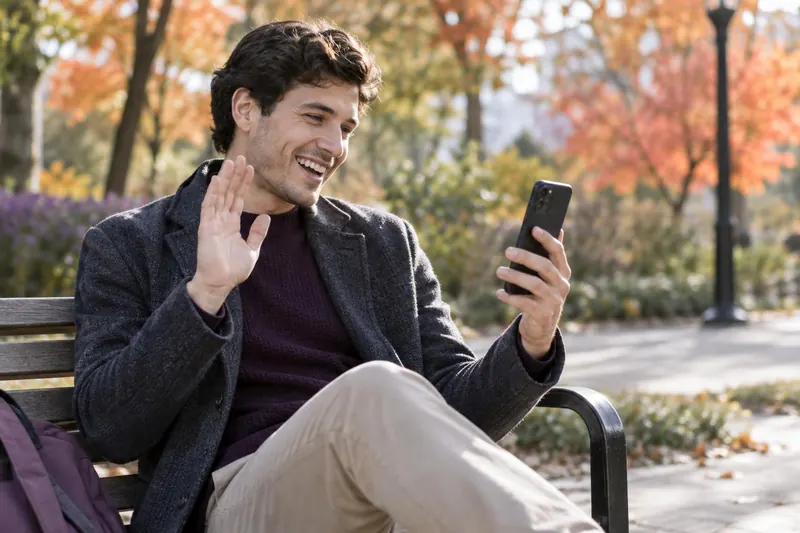 A young man on a park bench in autumn laughs and waves at his phone
